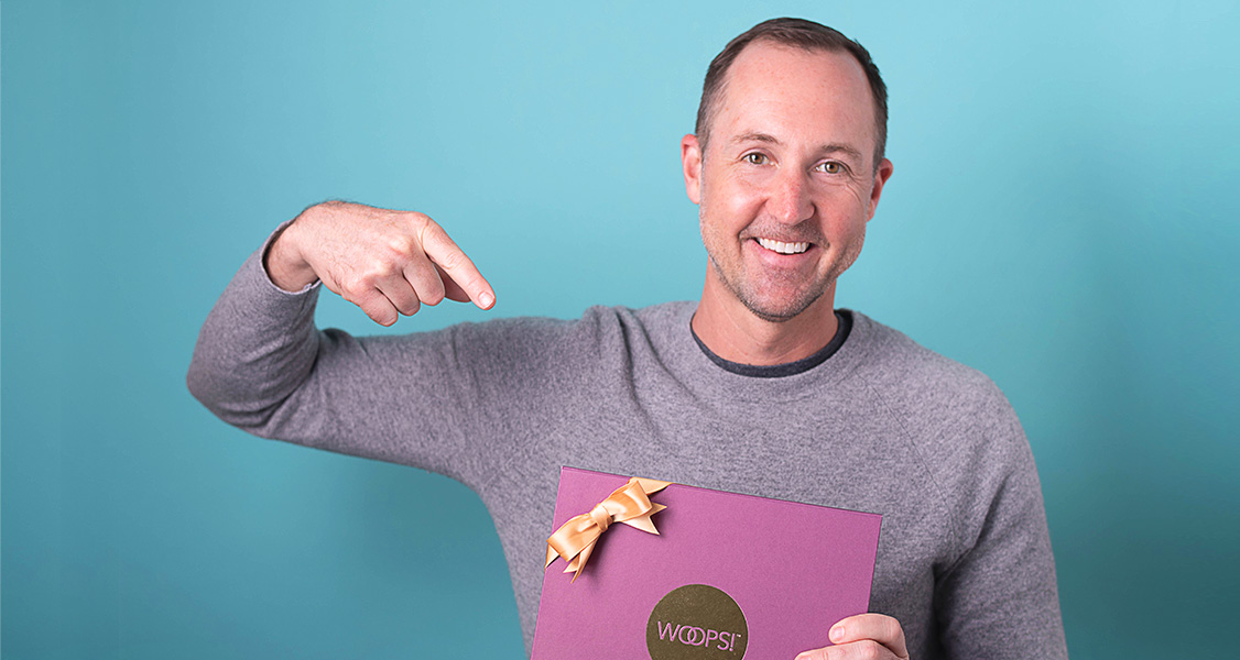 A smiling man is holding a purple French macaron box with a golden ribbon.