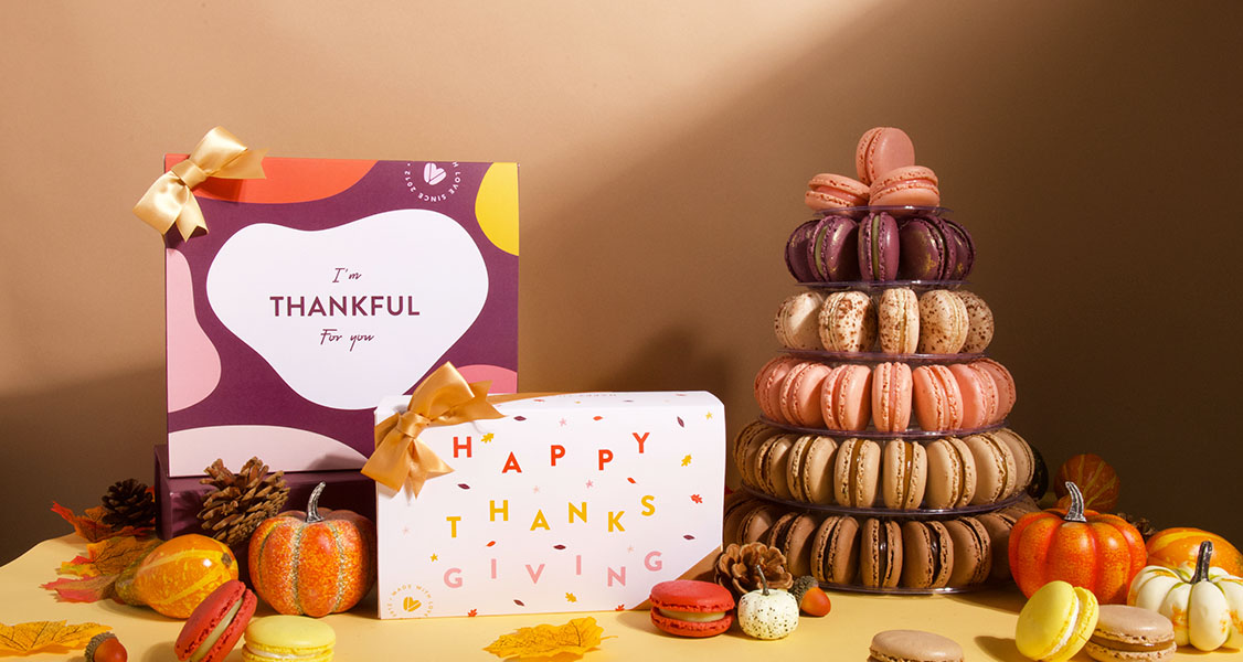 Two French macaron boxes with Thanksgiving sleeves and a French macaron pyramid are surrounded by assorted French macarons, leaves, and pumpkins.