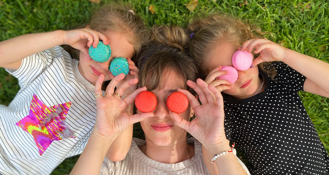 A smiling woman has two girls to her sides. All of them are holding French macarons to their eyes