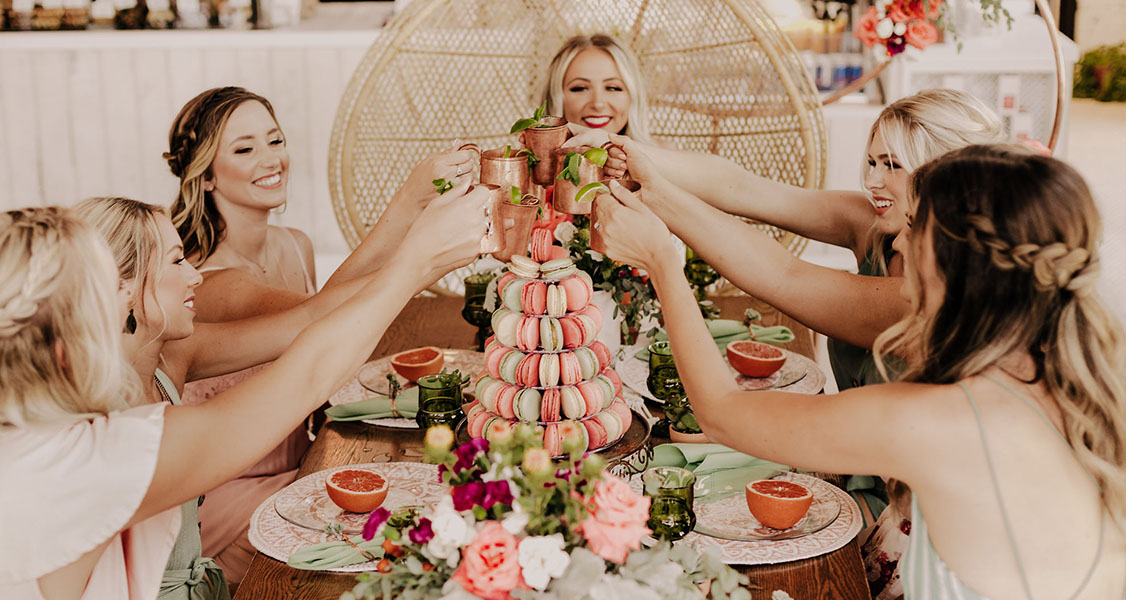 A decorated table has a French macaron pyramid at the center. Several women are toasting.