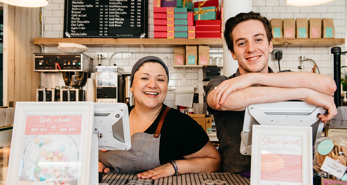 A smiling man and woman are behind a Woops! counter. Surrounding them are numerous French macaron boxes and pastries.