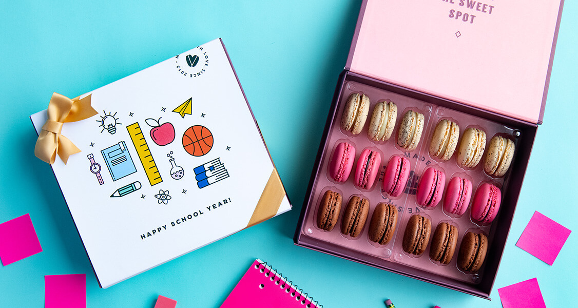 A box full of assorted French macarons has a French macaron box with a Back to School sleeve to its left. Below both boxes are some pink post-it notes, a notebook, and an eraser.