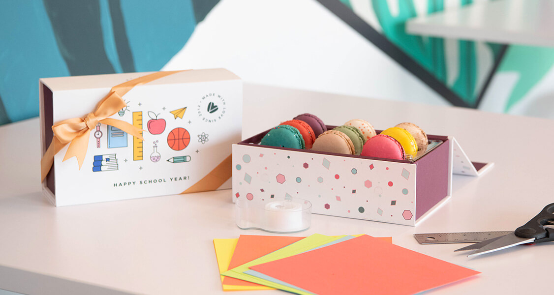 A box full of assorted French macarons has a French macaron box with a Back to School sleeve to its left. To the right are some post-it notes, a ruler, and a black scissor.