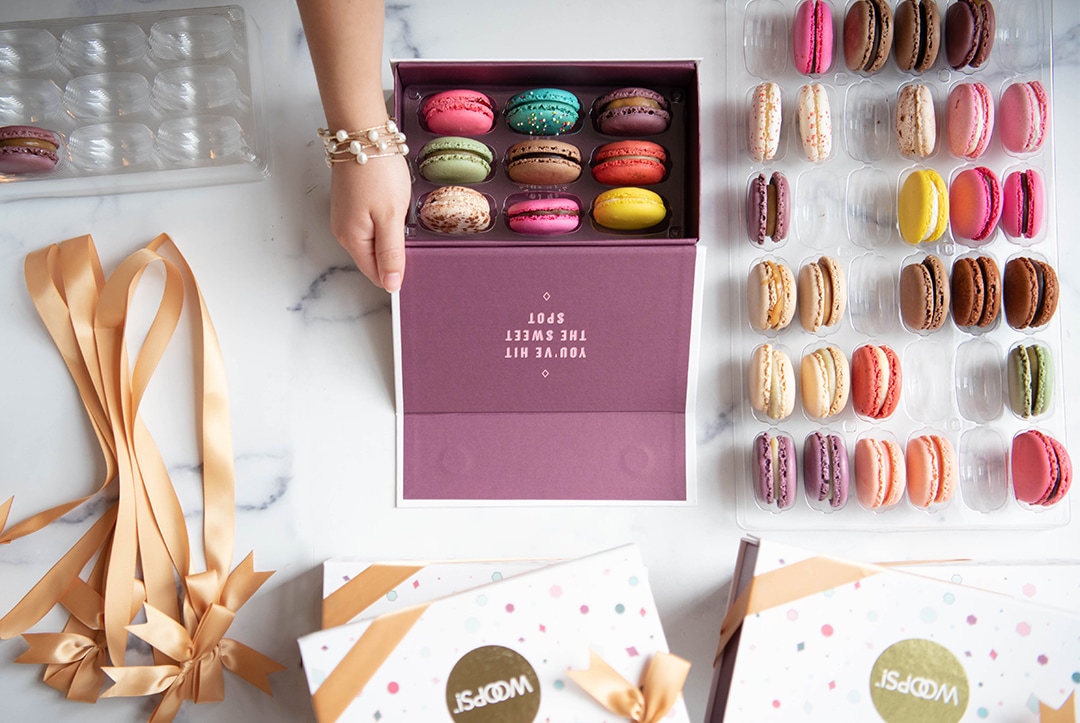 One hand is organizing a Woops! box filled with assorted macarons, some boxes, trays, and ribbons lying around