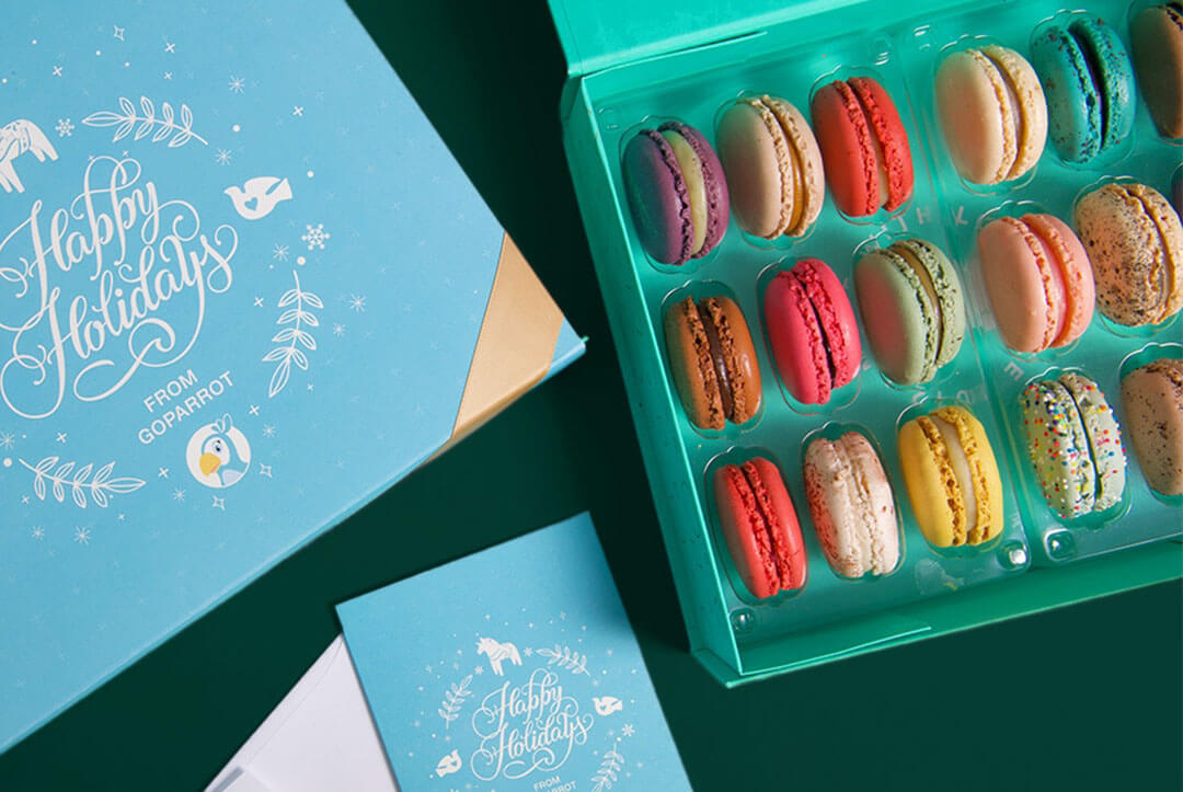 A box full of assorted French macarons has a French macaron box with a customized holiday sleeve and a customized greeting card to its left