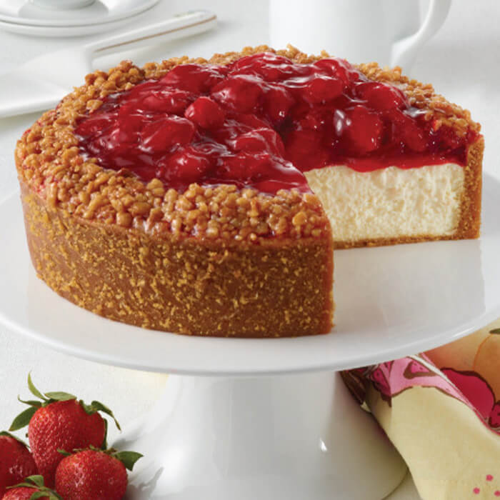 A strawberry cheesecake is on top of a white tier and surrounded by strawberries and white crockery.
