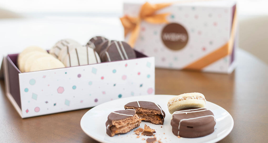 A plate full of assorted alfajores has a box of alfajores to its left and a French macaron box behind.