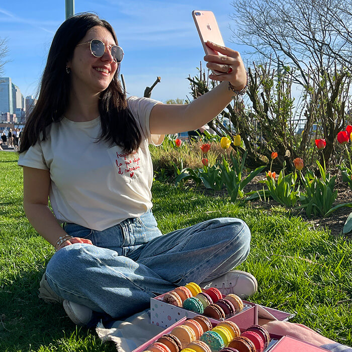 smiling woman that’s taking a selfie has two boxes full of assorted French macarons in front of her.
