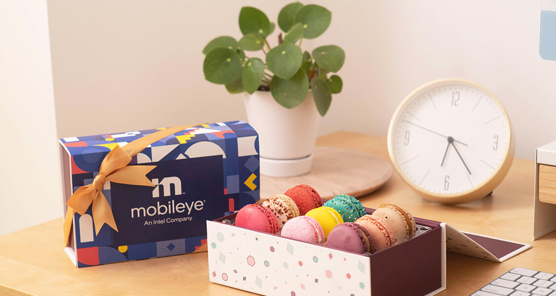 A box full of assorted French macarons has a French macaron box with a custom sleeve to its left and a plant and clock behind