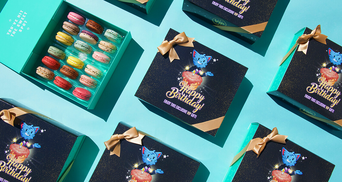 A box full of assorted French macarons is surrounded by French macaron boxes with custom "Happy Birthday" sleeves.