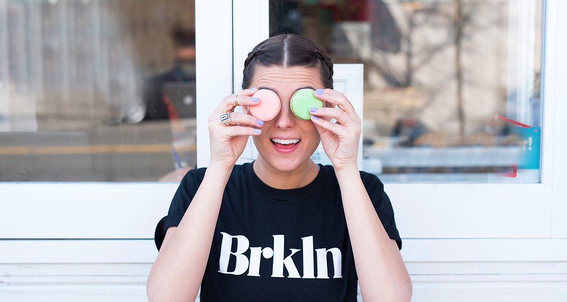 A smiling woman is holding 2 French macarons to her eyes.