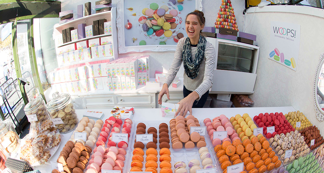 A smiling woman has a counter full of assorted French macarons in front of her.