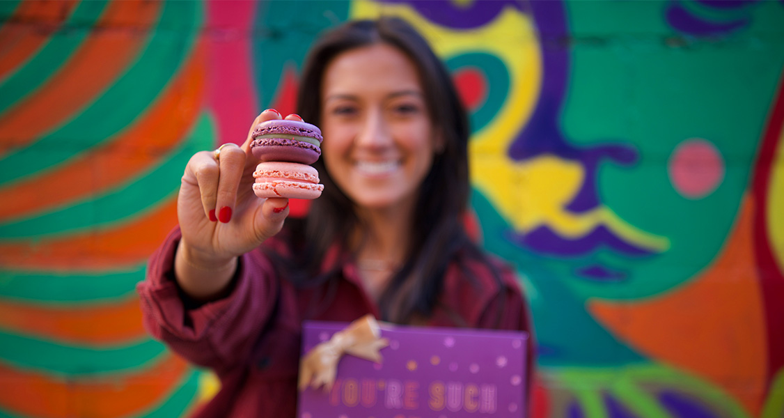 A smiling woman is holding a Woops! macaron box in one hand and two French macarons in the other.