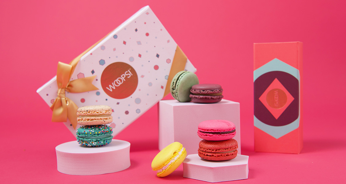 Several French macarons have two Woops! macaron boxes behind them.