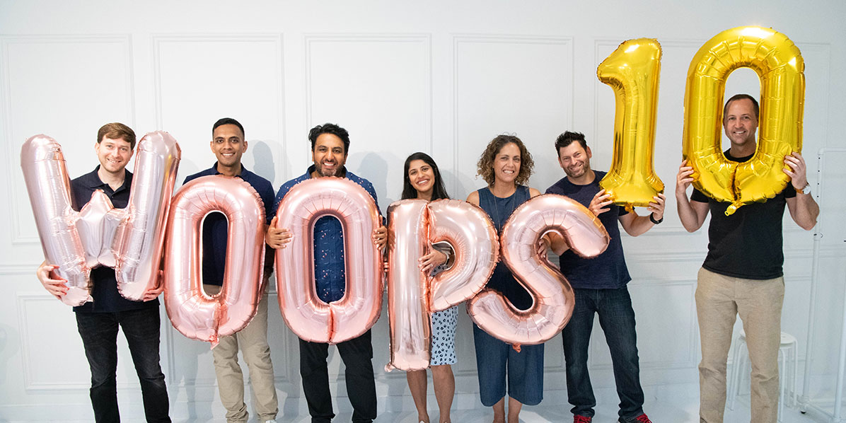 5 men and 2 women are holding pink and yellow balloons that spell Woops 10.