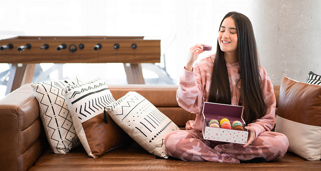 A smiling woman in pajamas is sitting on a brown couch while holding a box of 9 assorted French macarons.