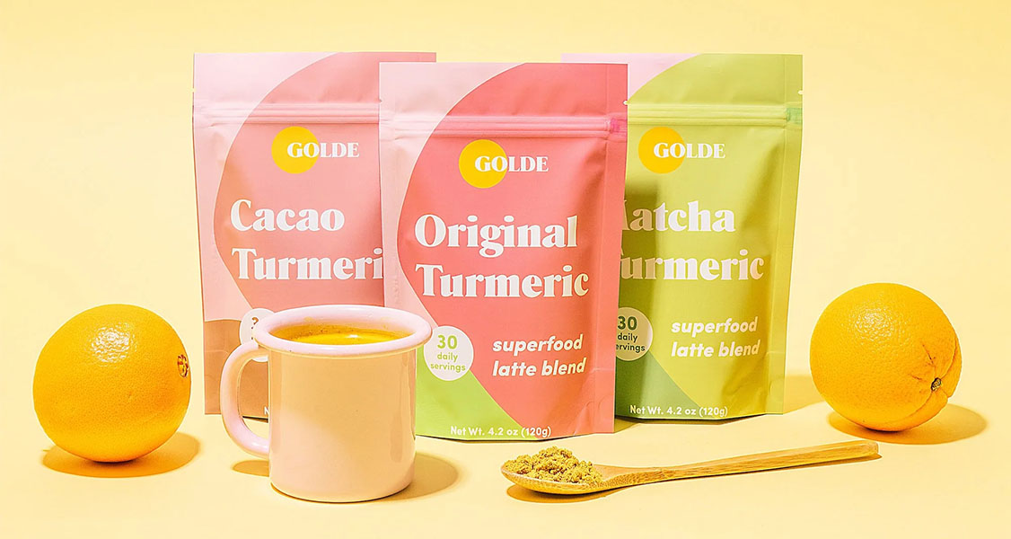 Three bags filled with wellness drinks are surrounded by oranges, a wooden spoon with turmeric, and a mug filled with a yellow drink.