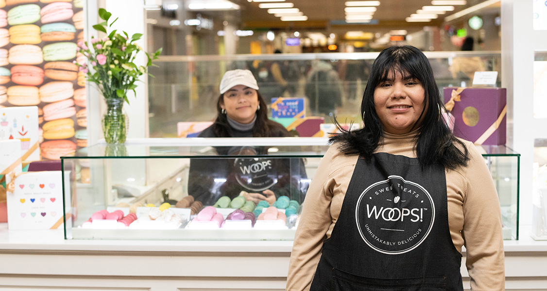 A smiling woman with a Woops! apron is standing in front of a display counter full of assorted French macarons. Behind the counter is another smiling woman.