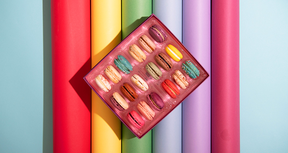 A box full of assorted French macarons has some colored cardboard rolls behind it.
