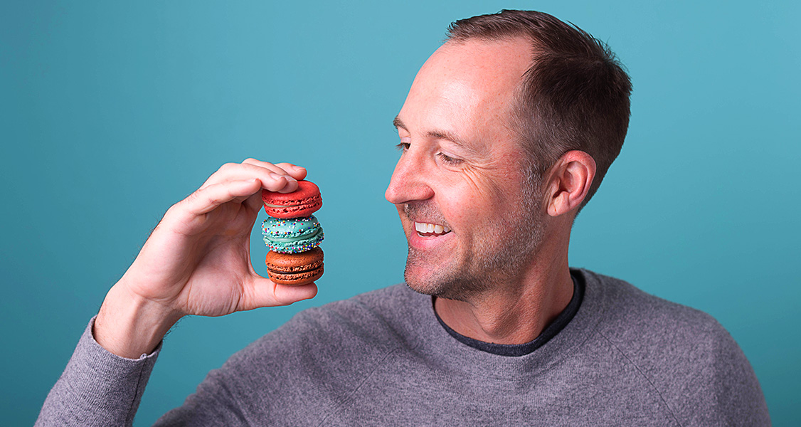 A smiling man with a gray sweater is holding 3 French macarons in one of his hands.