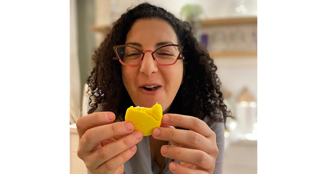 A smiling woman with curly hair is holding a Lemon Meringue French macaron in her hands.
