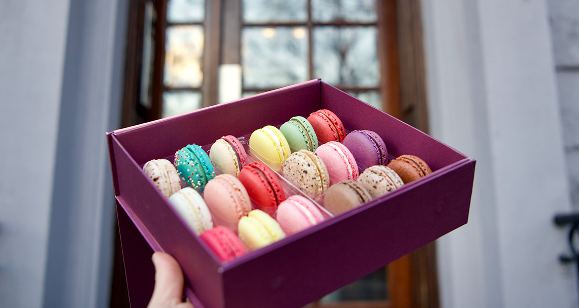 A hand is holding a box full of assorted French macarons in front of a doorstep.