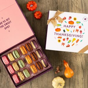 A box full of assorted macarons has a Woops! macaron box with a purple Thanksgiving sleeve to the left. Surrounding them are some paper leaves and tiny pumpkins.