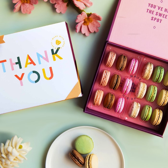 A green background features a Woops! box of 18 French macarons, a pink sleeve with white letters and a golden ribbon, some flowers, and a plate filled with macarons.A green background features a Woops! box of 18 French macarons, a pink sleeve with colored letters and a golden ribbon, some flowers, and a plate filled with macarons.