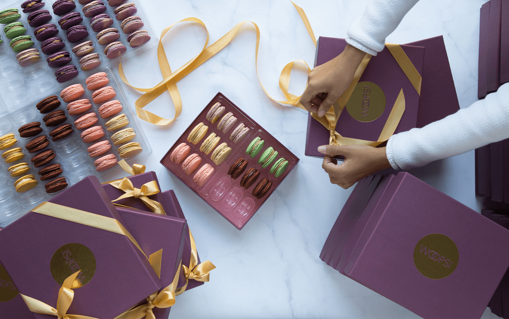 A box full of assorted French macarons is surrounded by several French macaron boxes with Holiday sleeves