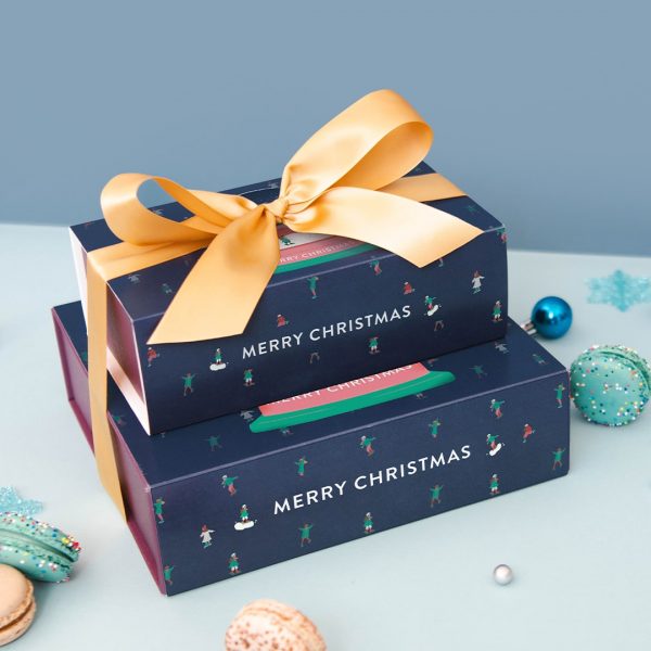 A stack of two French macaron boxes tied together by a golden ribbon is surrounded by assorted macarons and holiday ornaments.