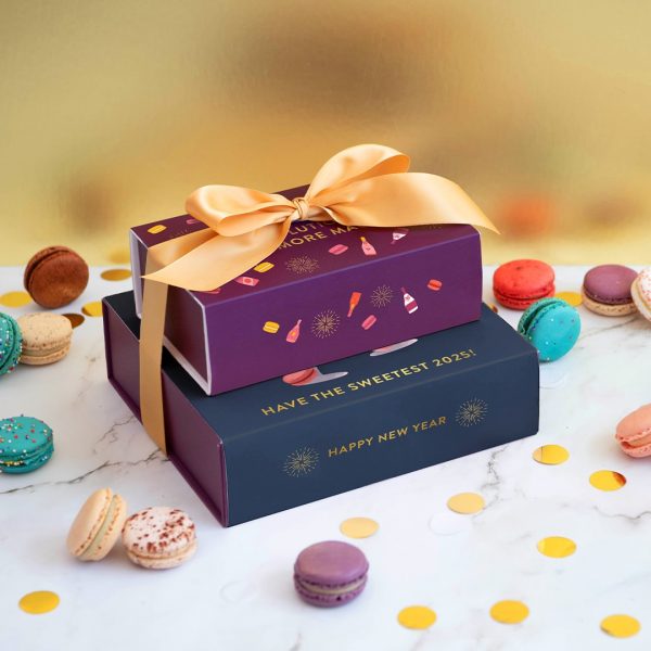 A stack of two French macaron boxes with New Year's sleeves surrounded by assorted macarons and confetti.