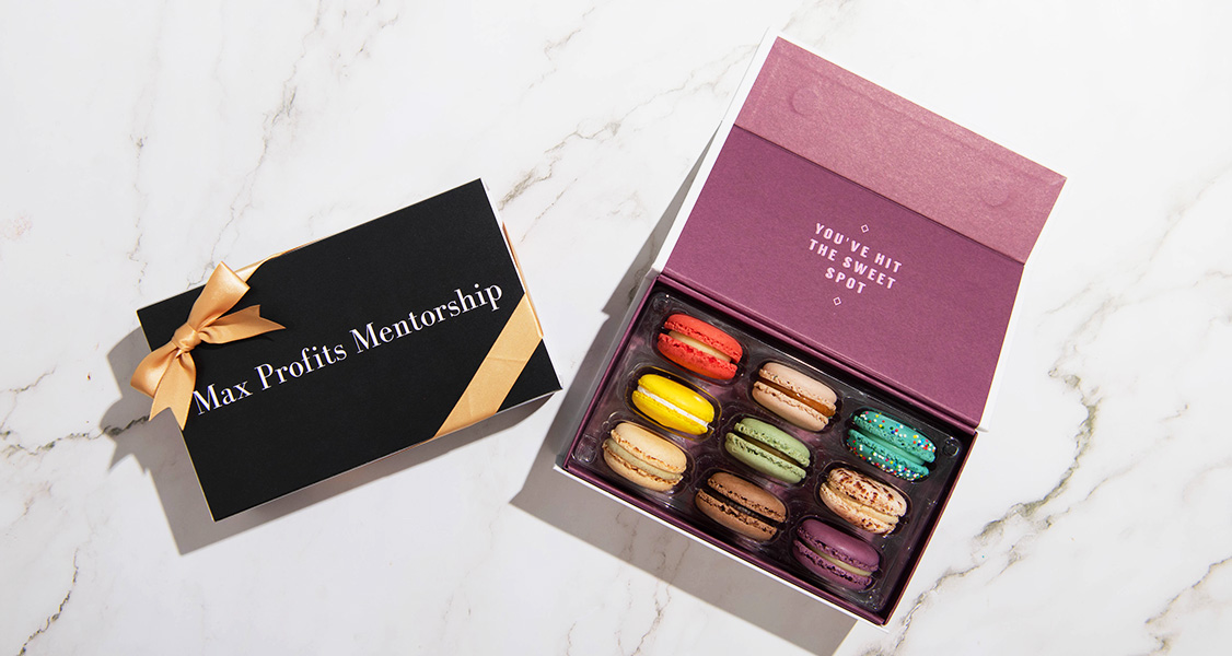A box full of assorted French macarons is surrounded by French macaron boxes with custom sleeves and golden ribbons.