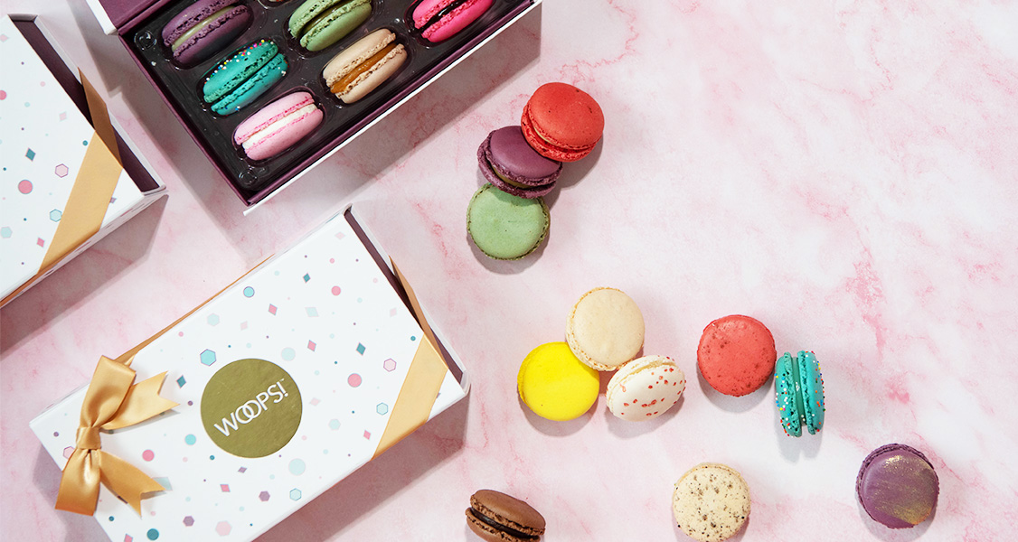 A box full of assorted French macarons has 2 French macaron boxes to the left. Lying around are some assorted French macarons.