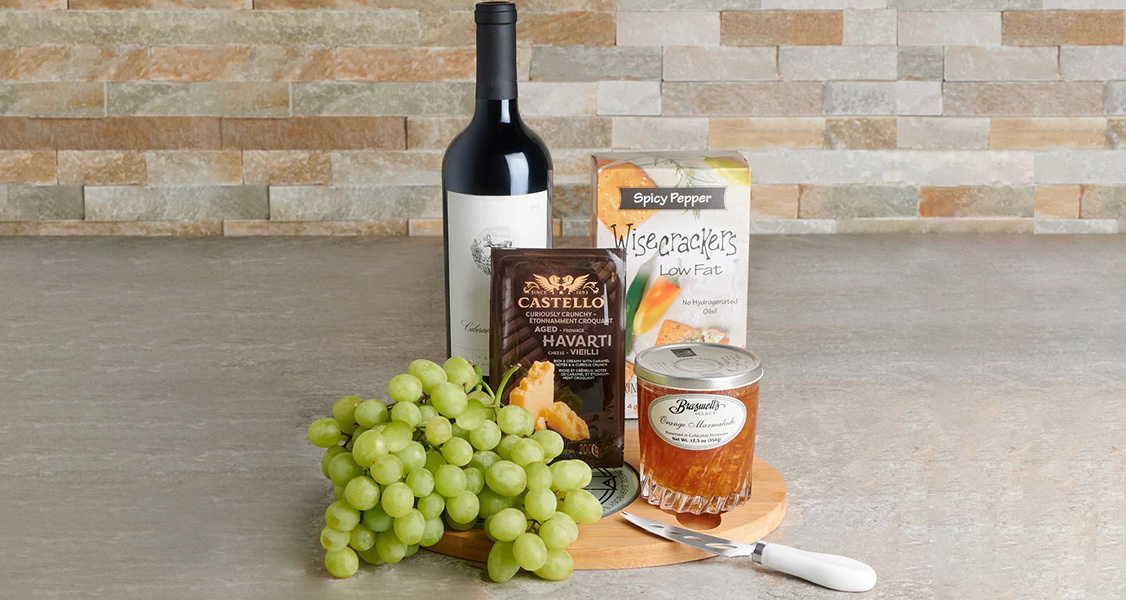 A bottle of wine, spicy pepper crackers, aged Havarti cheese, orange marmalade, fresh green grapes, and a wood & glass cutting board with a cheese knife in it.
