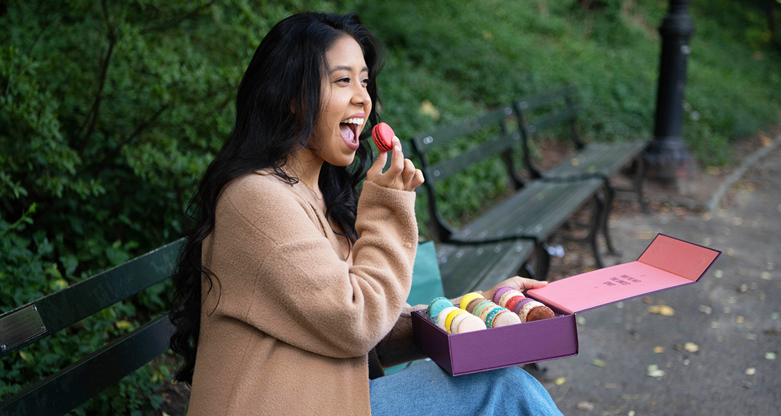A smiling woman is eating French macarons while sitting on a bench.