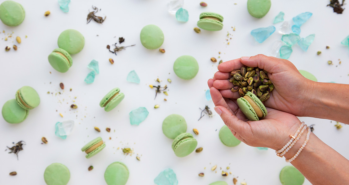 Two female hands are holding a Pistachio macaron and some pistachios. Below are numerous Pistachio French macarons, pistachios, and blue rocks.