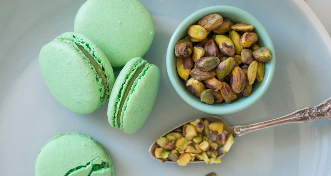 4 Pistachio French macarons have a spoon and a tiny bowl full of pistachios to the right.