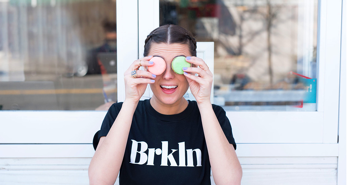 A smiling woman is holding two French macarons to her eyes.