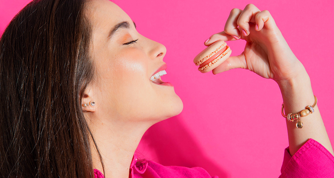 A smiling woman with a pink shirt is holding a Rose French macaron.