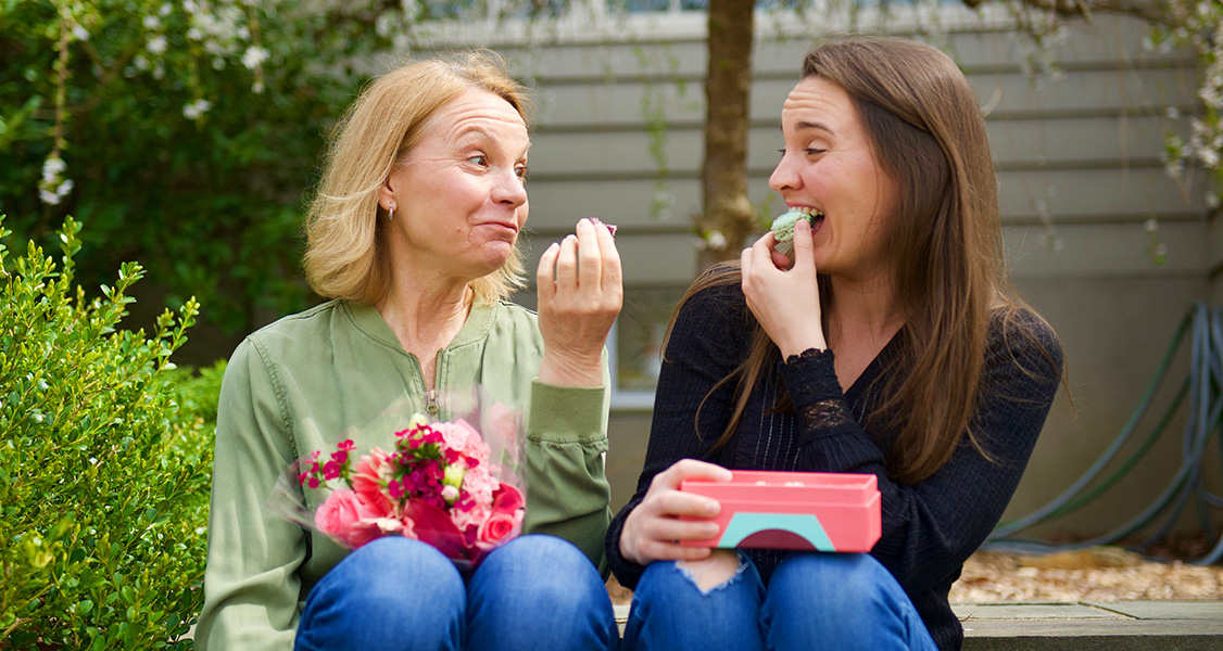 Two smiling women are sitting and looking at each other while holding French macarons in their hands.