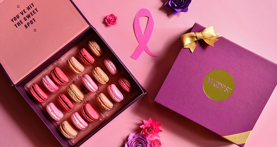A box full of assorted French macarons has a purple French macaron box, some flowers, and a BCA ribbon to its right.