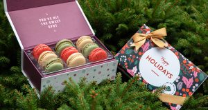 A box full of assorted French macarons has a French macaron box with a holiday sleeve and a golden ribbon to the right.