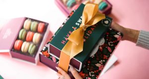 Two hands are holding a French macaron stack. On the background is a box full of assorted French macarons.