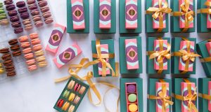 Numerous pink macaron favor boxes with golden ribbons have two trays full of assorted macarons and a box with French macarons to the left.