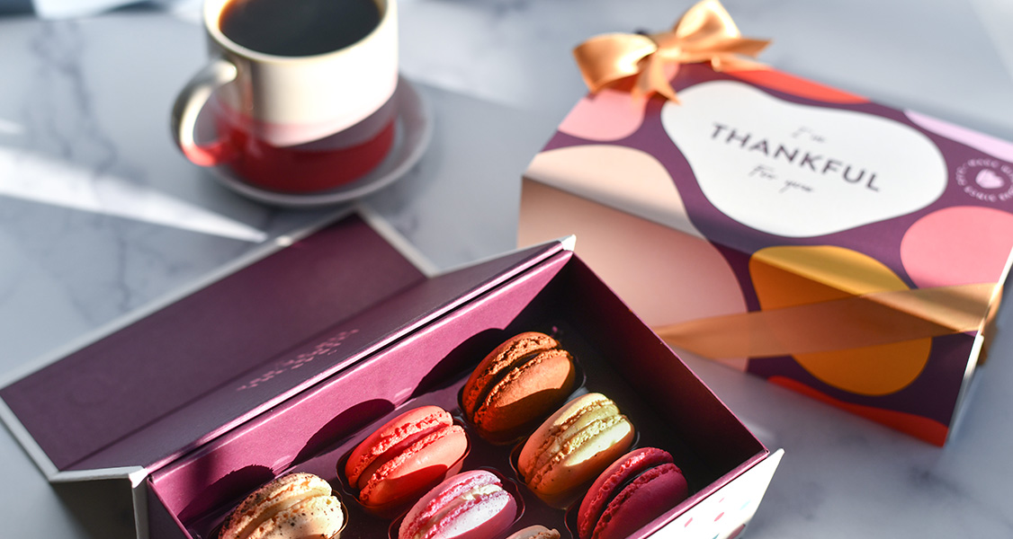 A box full of assorted French macarons has a French macaron box with a Thanksgiving sleeve to the right and a cup of coffee above.