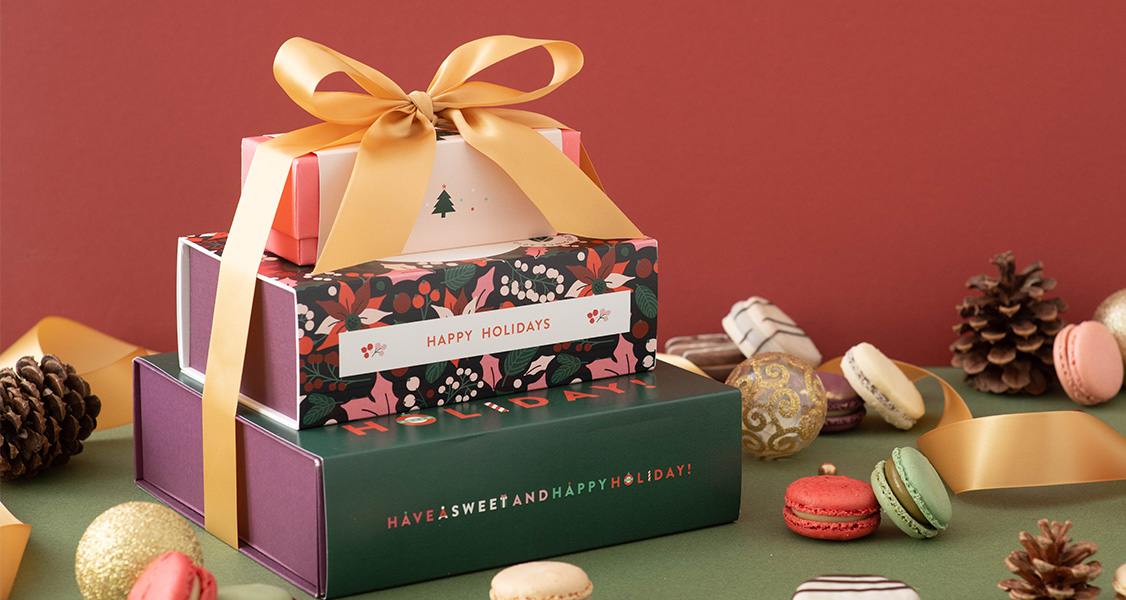 Three French macaron boxes are stacked and tied with a golden ribbon. Surrounding the stack are some assorted French macarons, alfajores, and holiday ornaments.