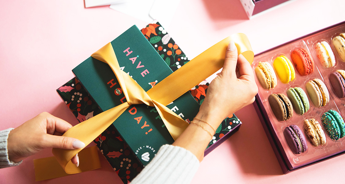 Two female hands are tying a golden ribbon on top of a French macaron stack. To the right are two boxes full of assorted French macarons.