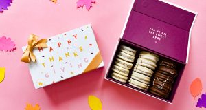 A box full of assorted alfajores has a French macaron box with a Thanksgiving sleeve to the left. Surrounding them are numerous paper leaves.