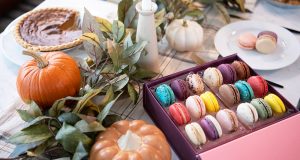 A box full of assorted French macarons and a plate with three French macarons are surrounded by pumpkins, decorative leaves, a candle, and a pie.