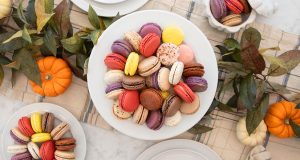 Four plates full of assorted French macarons are surrounded by pumpkins and leaves.